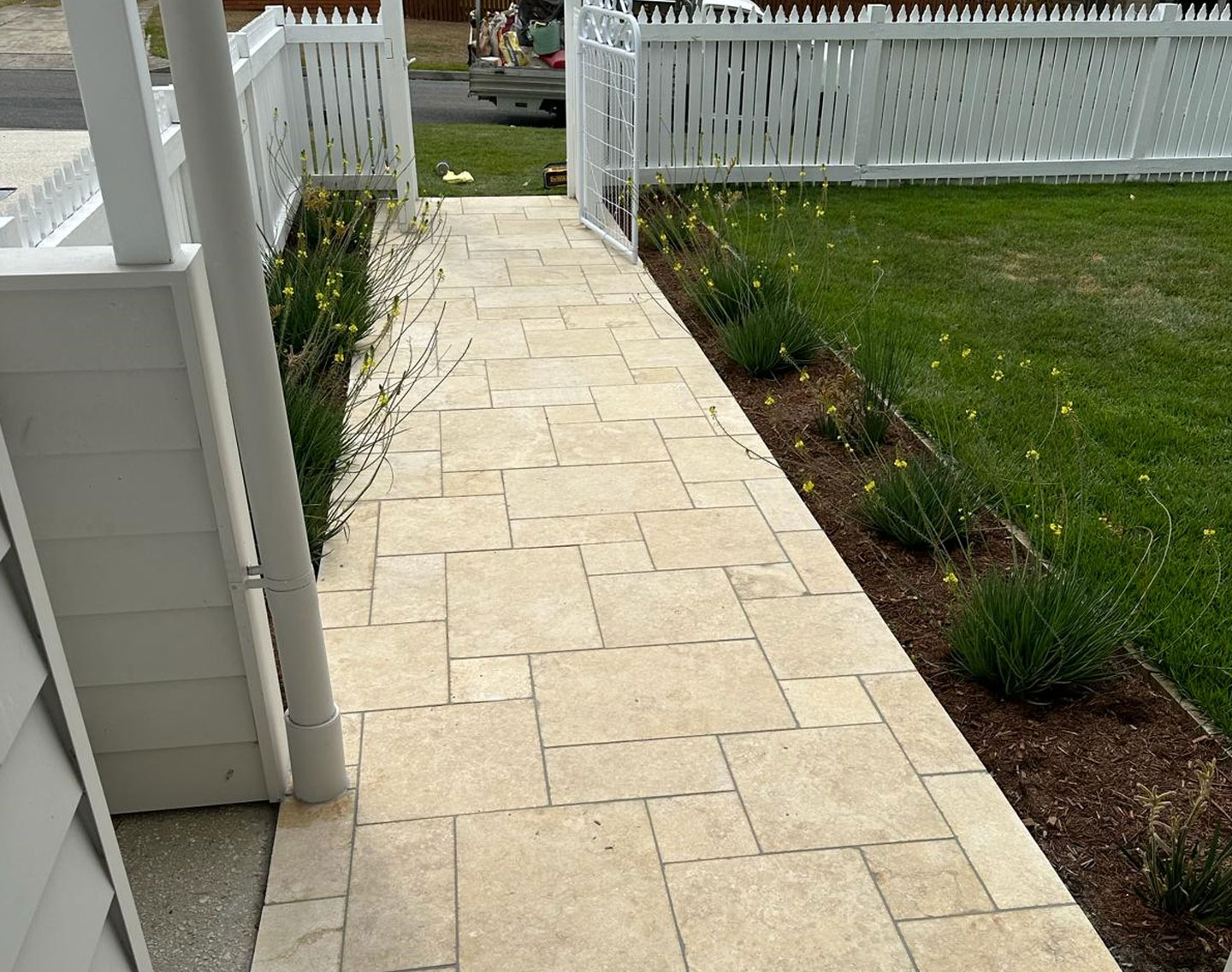 SOLUTIONS-FOR-ALL-YOUR-PAVING-AND-TILING-NEEDS Beige paved path running beside a verandah and lawn to a white picket fence