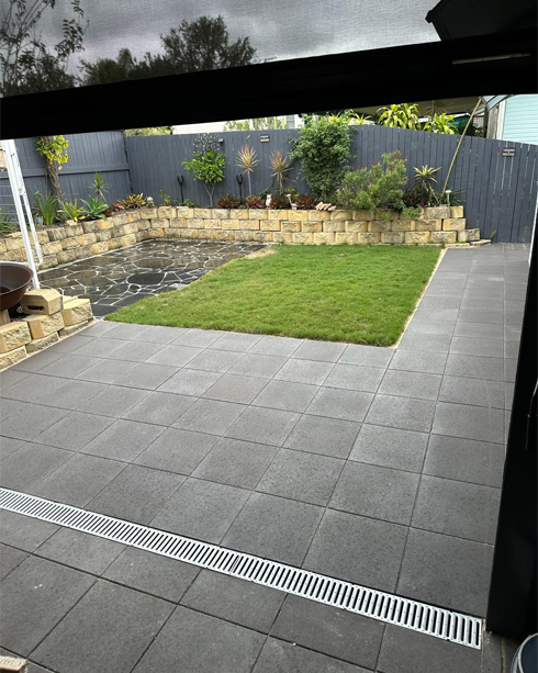 Charcoal paved backyard patio with a drainage grate, lawn and block retaining wall